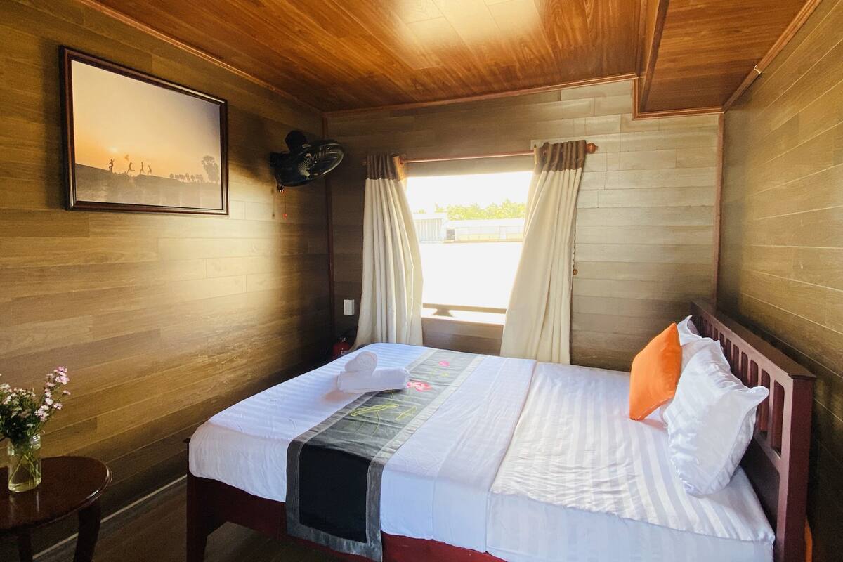 Ein Zimmer mit Holzwänden, einem Bett, einem kleinen Tisch mit Blumenvase und einem gerahmten Bild an der Wand.