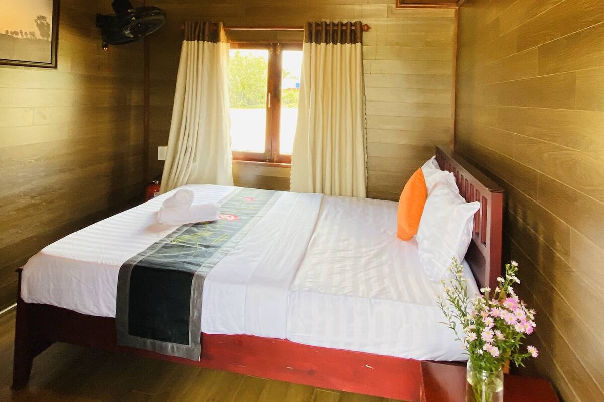 Ein Zimmer mit Holzvertäfelung, einem Bett, einem Fenster mit Vorhängen, einem Wandventilator und einem gerahmten Bild.