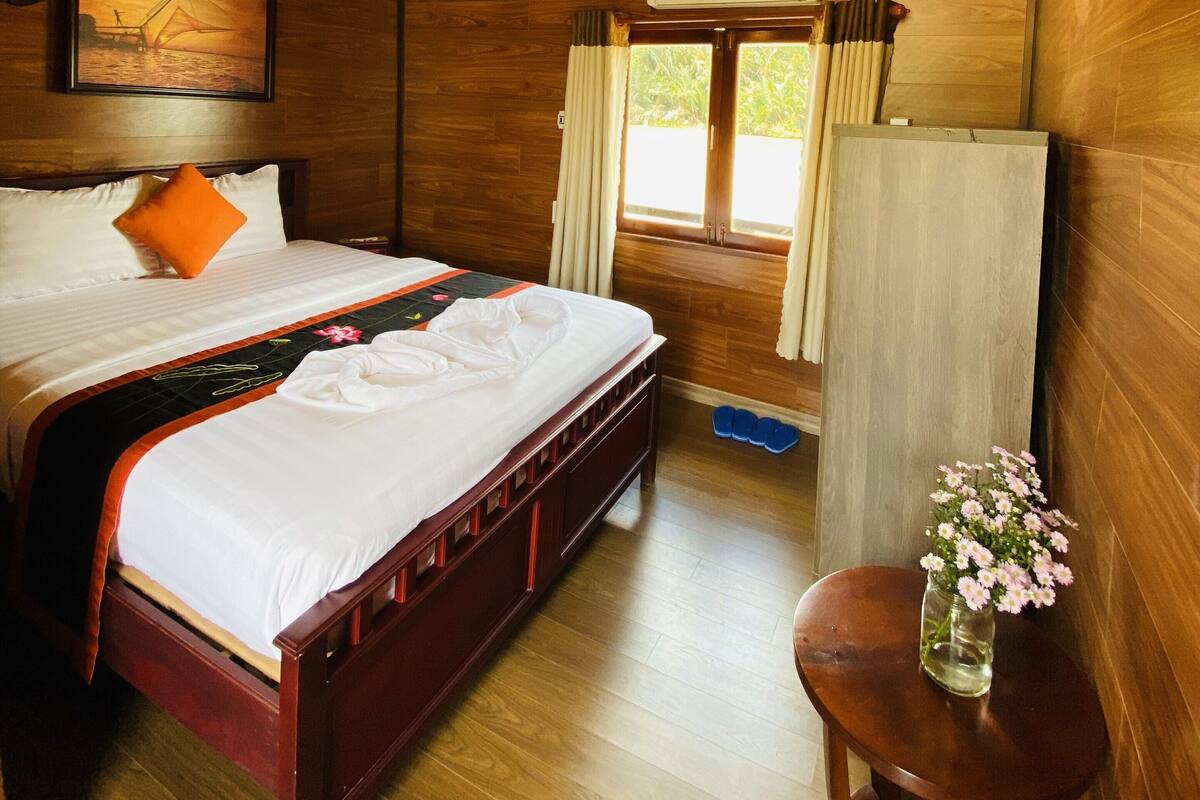 Ein Zimmer mit Holzboden, einem Bett, einem Standventilator, einem Fenster mit Vorhängen und einem kleinen Tisch mit einer Vase.