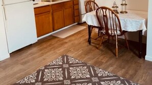 A compact kitchen with wooden cabinets, a white refrigerator, and a stove. There is a dining area with two wooden chairs and a table, and a patterned rug on the floor. - North Star Place Apt 8 (Fairbanks)