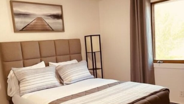 A bedroom with a large bed, a headboard, a bedside table, a floor lamp, a window with curtains, and a painting on the wall. - North Star Place Apt 8 (Fairbanks)