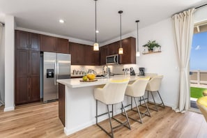 A modern kitchen with a central island, bar stools, wooden cabinets, and a view of the outdoors. - @ Marbella Lane - Stylish House + Impressive Views (Waianae)