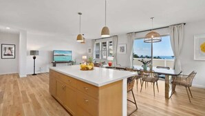 Cozinha moderna com ilha central, armários de madeira, iluminação pendente e área de jantar com cadeiras de vime.