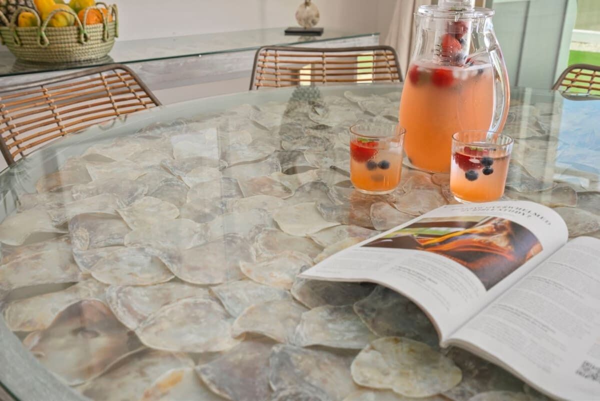 Una mesa redonda de vidrio con un diseño de mosaico, una jarrón de vidrio con una bebida, dos vasos con fruta y una revista abierta.