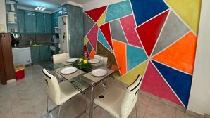 A dining area with a glass table and chairs, a colorful geometric wall mural, and a kitchen in the background.