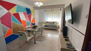 A dining area with a glass table, chairs, and a colorful geometric wall. A sofa and a TV are in the background.