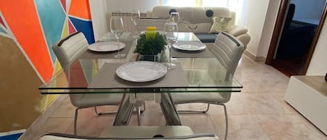 A dining area with a glass-top table, white chairs with black bows, and a colorful geometric wall.