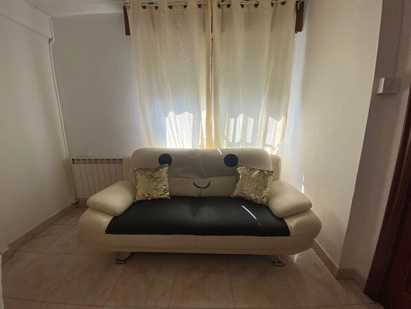 Un sofá beige con dos almohadones decorativos, un radiador y una ventana con cortinas traslúcidas.