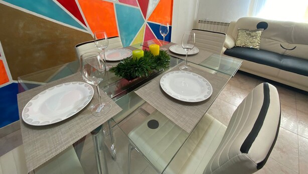 A dining table set with plates, glasses, and candles, surrounded by a colorful geometric wall and a sofa with a cushion.