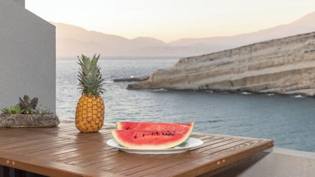 Une table en bois sur laquelle sont posés un ananas et une tranche de pastèque, le tout sur une assiette. La table est placée de manière à offrir une vue sur un paysage côtier, avec des falaises et la mer.