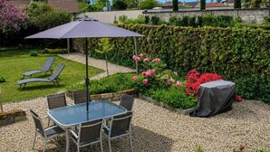 Eine Terrasse mit Tisch und Stühlen, einem schattenspendenden Sonnenschirm und einem Garten mit Blumen und einer Hecke.