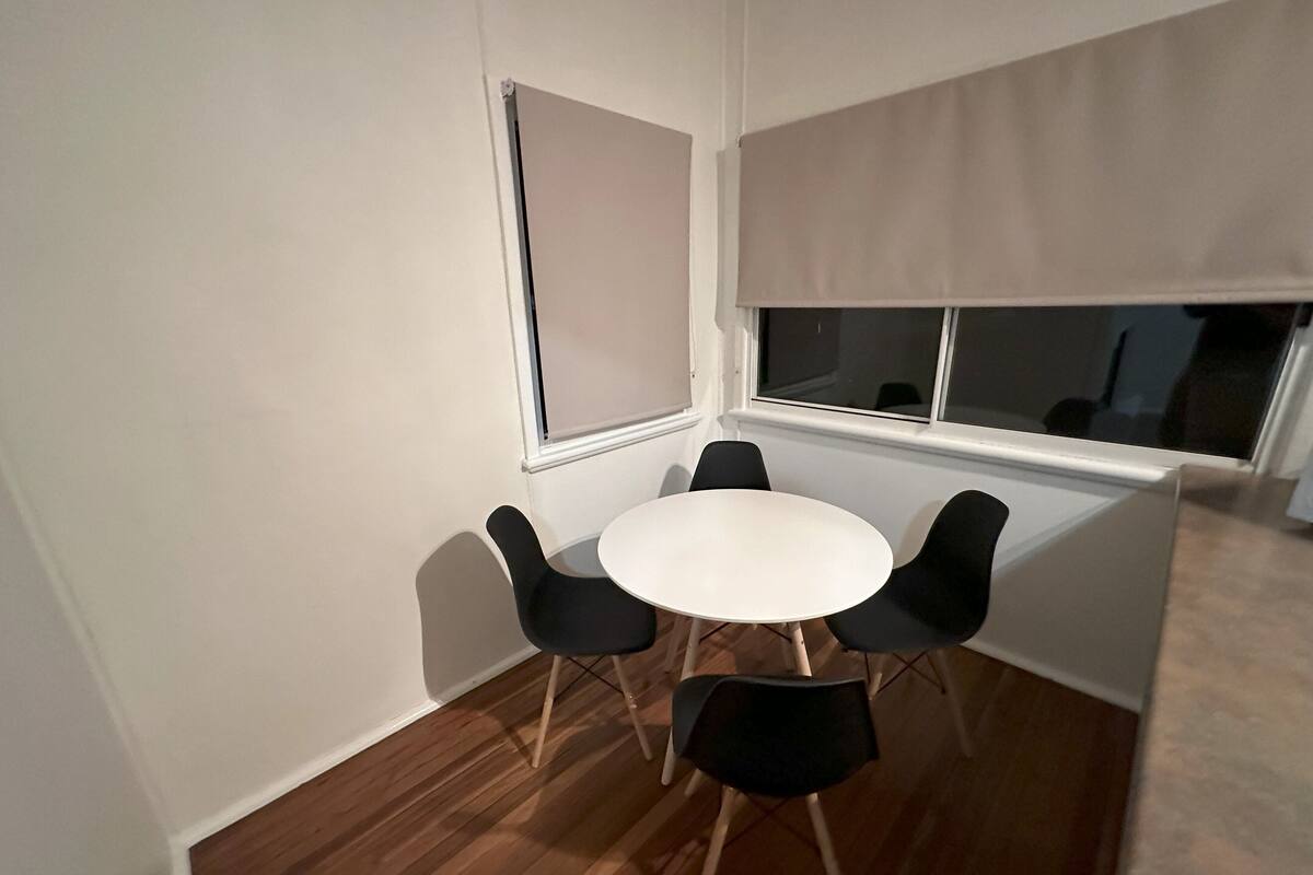 Une petite pièce avec une table ronde et quatre chaises, une fenêtre avec des stores et un plancher en bois.
