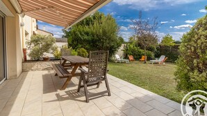 Une terrasse avec une table et des chaises en bois, un espace couvert et une cour arrière verdoyante avec des fauteuils de jardin.