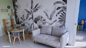 Uma sala de estar com um sofá cinzento, uma mesa redonda com cadeiras, um tapete tecido e uma parede com padrão de folhagem tropical.