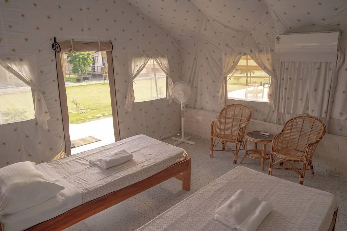 Quarto estilo tenda com duas camas, um ventilador, cadeiras de vime e vista para o exterior.