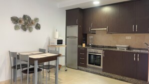 A modern kitchen with dark wooden cabinets, a stainless steel refrigerator, a microwave, and a built-in oven. There is a dining table with chairs and a small island with a sink and a blender.