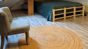 A loft bedroom with a sloped ceiling, a bed with a green bedspread, a wooden bench, a woven rug, and a window with curtains. - La Maison du Moulin (Saint-Georges-sur-Eure)