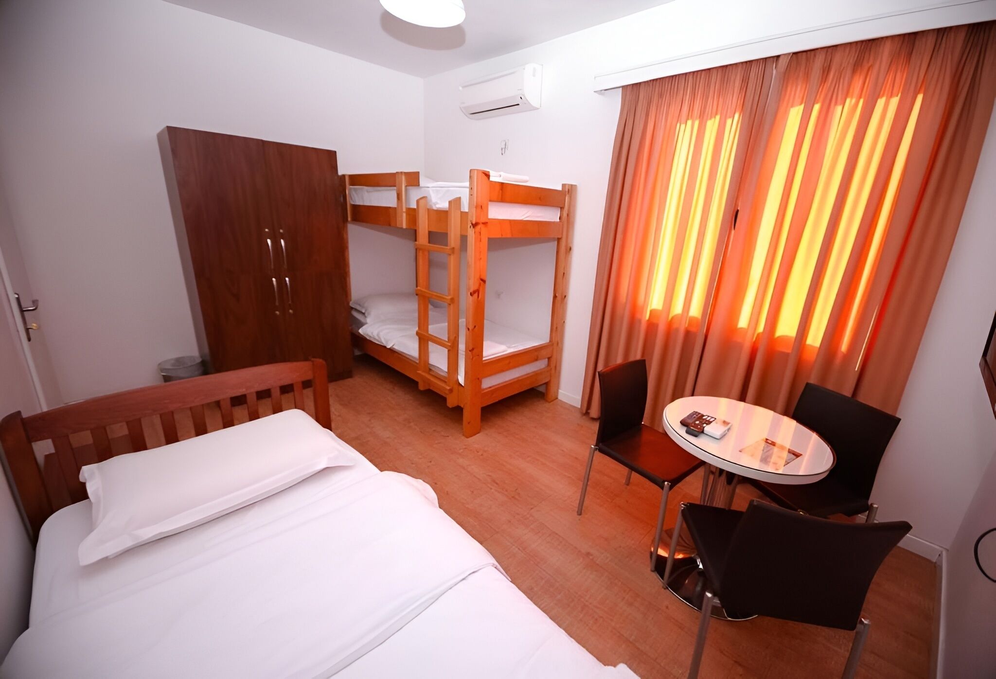 Een hotelkamer met stapelbedden, een houten garderobe, een klein rond tafeltje met stoelen en een raam met gordijnen.