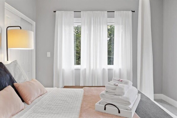 A bedroom with a bed, a bedside lamp, a window with white curtains, and a stack of towels on a tray.