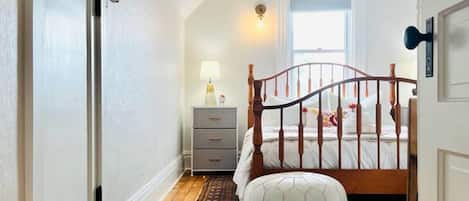 A bedroom with a wooden floor, a bed with a wooden headboard, a nightstand, a lamp, and a window.