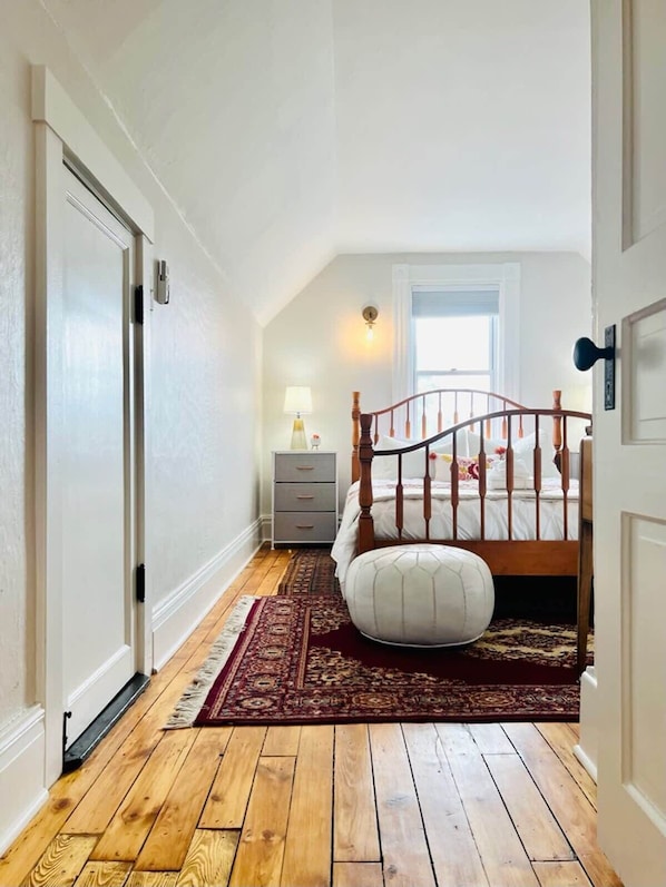 A bedroom with a wooden floor, a bed with a wooden headboard, a nightstand, a lamp, and a window.