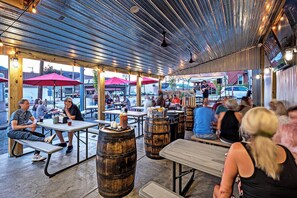 An outdoor cafe with wooden tables and barrels, featuring a corrugated metal roof and red umbrellas.