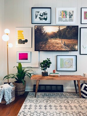 A living room with a large TV, framed artwork on the wall, a wooden coffee table, and a potted plant.