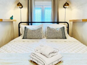 A neatly made bed with a white quilt and two striped pillows, a stack of white towels, and two wall-mounted lamps.