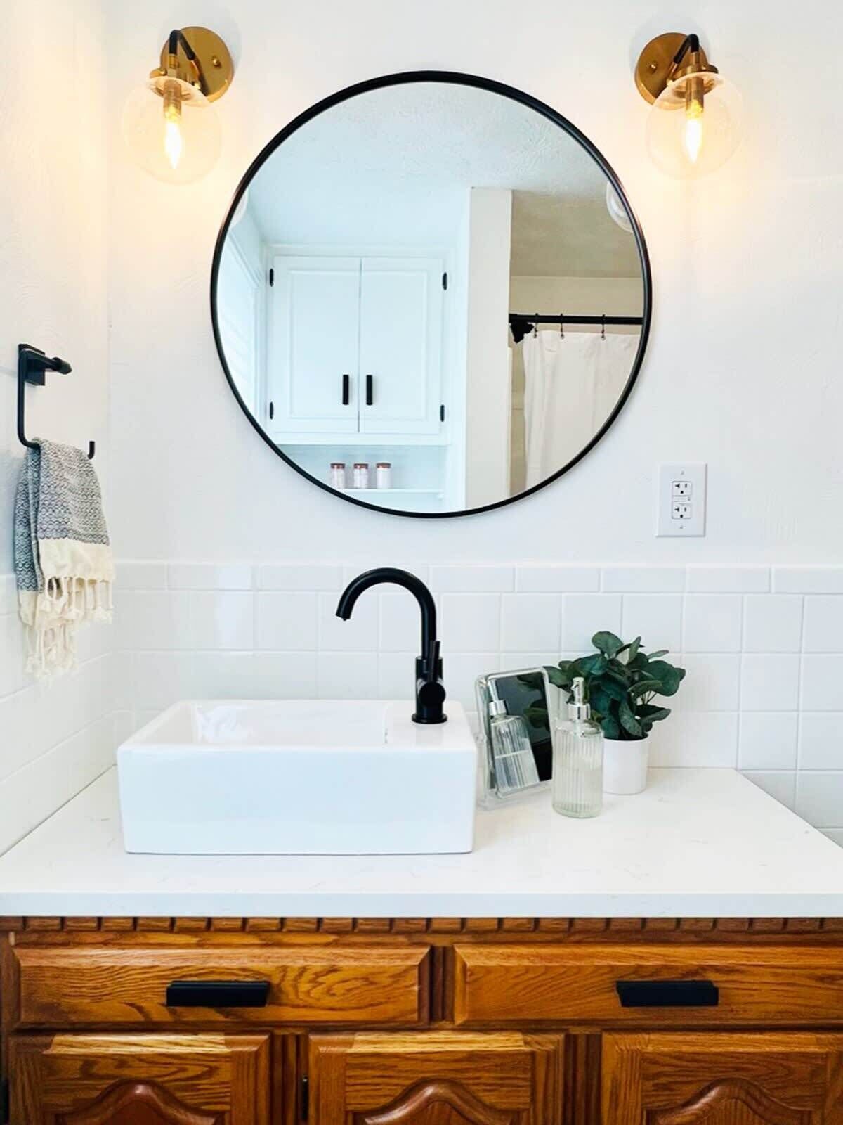 Une salle de bain avec un lavabo blanc, un robinet noir et un miroir rond au-dessus. On y trouve deux lampes fixées au mur et un porte-serviettes avec une serviette.