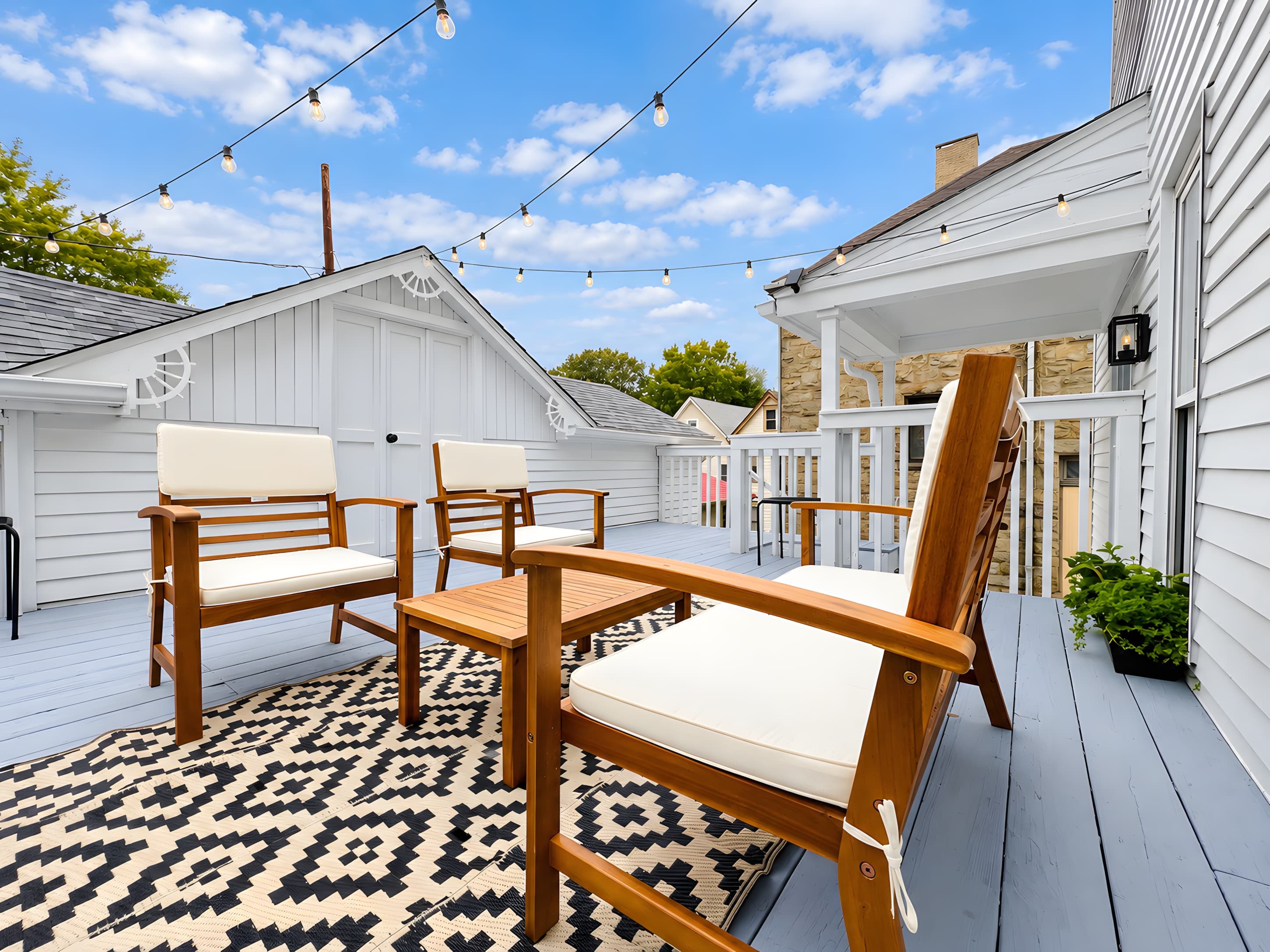 A deck with wooden furniture and string lights, white siding, and a patterned rug.