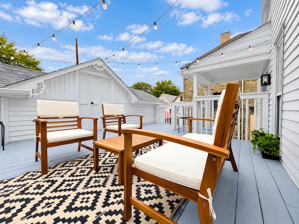 A deck with wooden furniture and string lights, white siding, and a patterned rug.