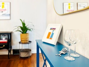 A room with a blue table, a framed chart, two wine glasses, a plant, and a small side table with a basket.
