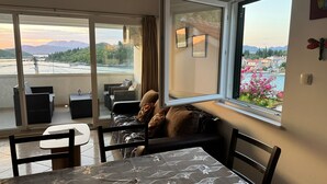 Une pièce comprenant une table à manger, des chaises, un canapé, une grande fenêtre donnant sur des montagnes et un plan d’eau, ainsi qu’un balcon avec du mobilier d’extérieur.