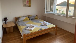 Une chambre à coucher avec un lit en bois, une table de chevet, une lampe, un tableau et une fenêtre donnant sur un plan d’eau et des collines.