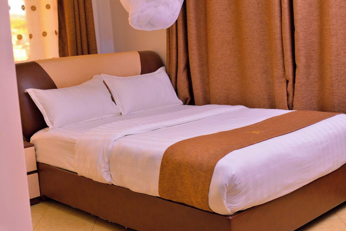 A neatly made bed with white linens and a brown accent, a nightstand with a lamp, and brown curtains.