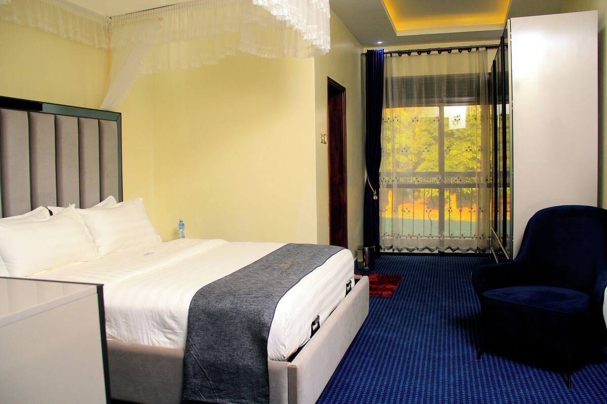A hotel room with a bed, a chair, a window with curtains, and a view of trees.