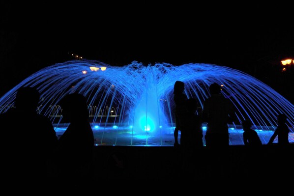 Una fuente con chorros de agua iluminados en azul, rodeada de siluetas de personas.