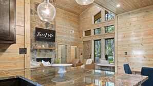 A spacious room with a large marble island, wooden walls, and a stone fireplace. - Pet-Friendly Forest Retreat for Families & Friends (Broken Bow)