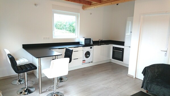 Ett kompakt kök med tvättmaskin, mikrovågsugn och en inbyggd ugn. Det finns två barstolar och ett bord.