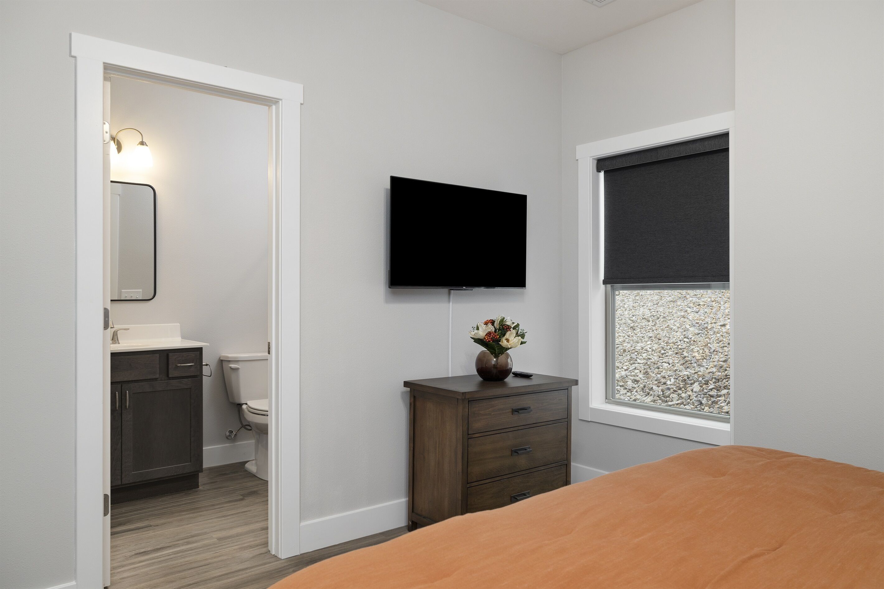 Un dormitorio con cama, cómoda, televisor montado en la pared y ventana con vista a un camino de grava.