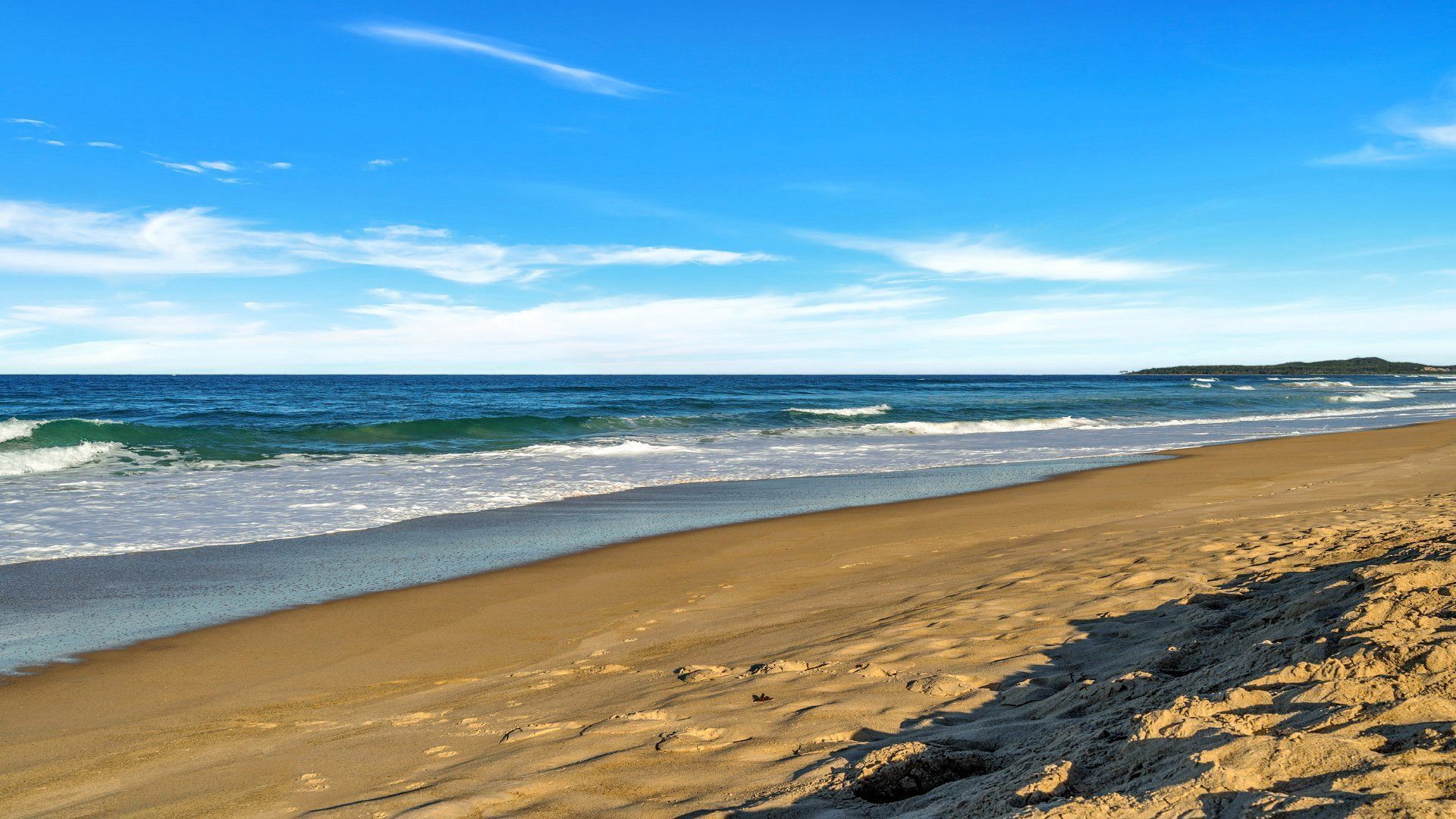 A beach with waves, a clear sky, and a distant landmass.