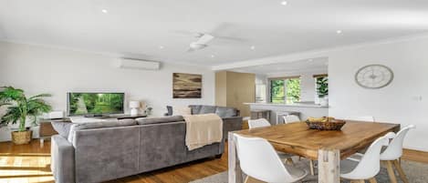A modern living room with a grey sofa, a wooden dining table, and white chairs.