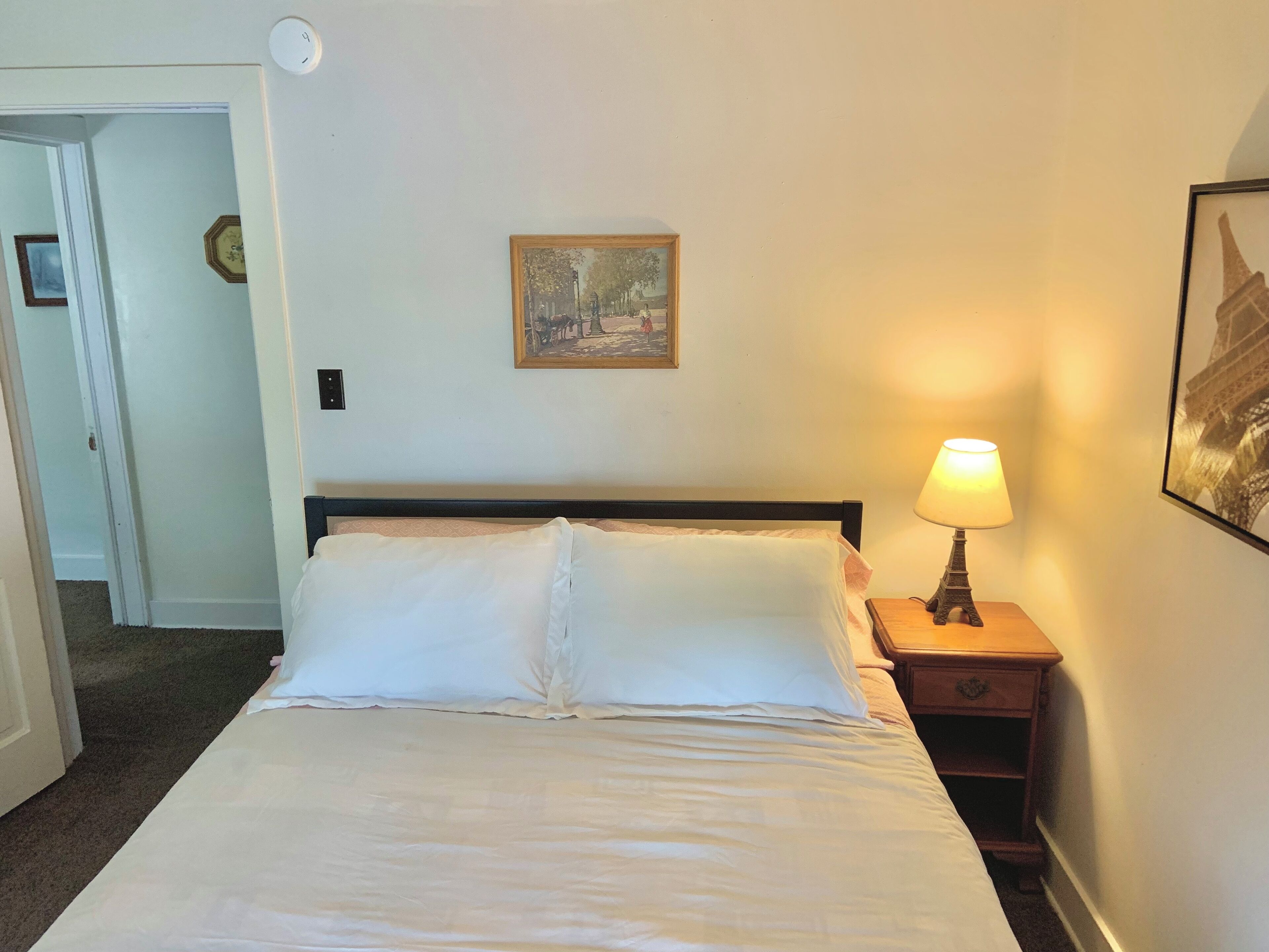 A neatly made bed with white linens, a bedside lamp, a small wooden nightstand, and a framed picture on the wall.