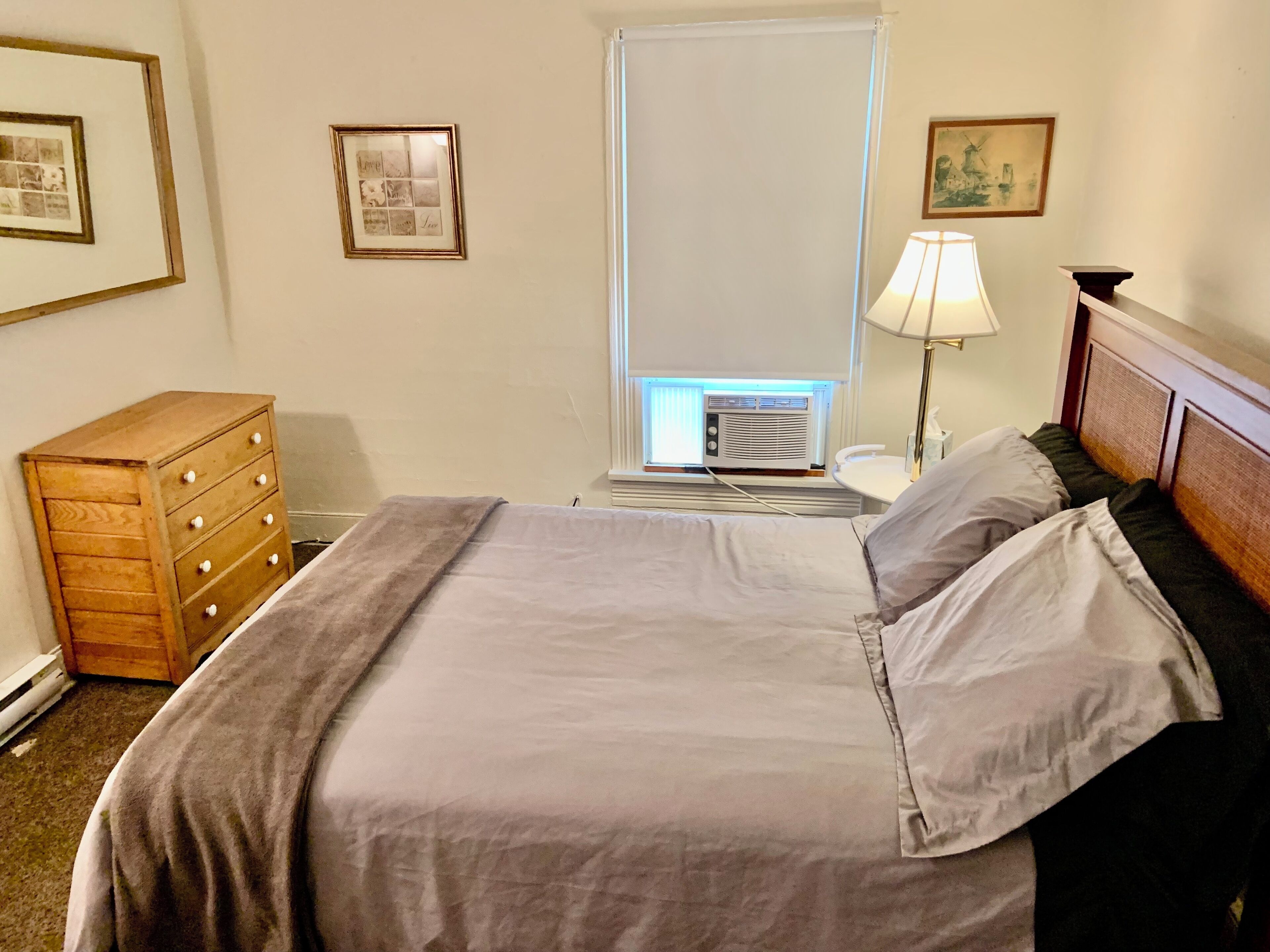 A neatly made bed with a gray blanket and pillows, a wooden dresser, a window with blinds, and a wall-mounted air conditioner.
