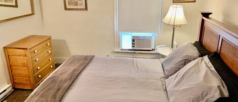 Una cama bien hecha con una manta gris y almohadas, un aparador de madera, una ventana con persianas y un aire acondicionado instalado en la pared.