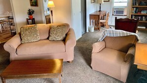 Un salón con un sofá beige, un sillón a juego, una mesa de centro de madera, un reloj de pared, una zona de comedor con una mesa y sillas, y una estantería.