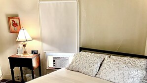 Una cama bien hecha con una manta escocesa, una mesita de noche con una lámpara, un reloj y un radiador.