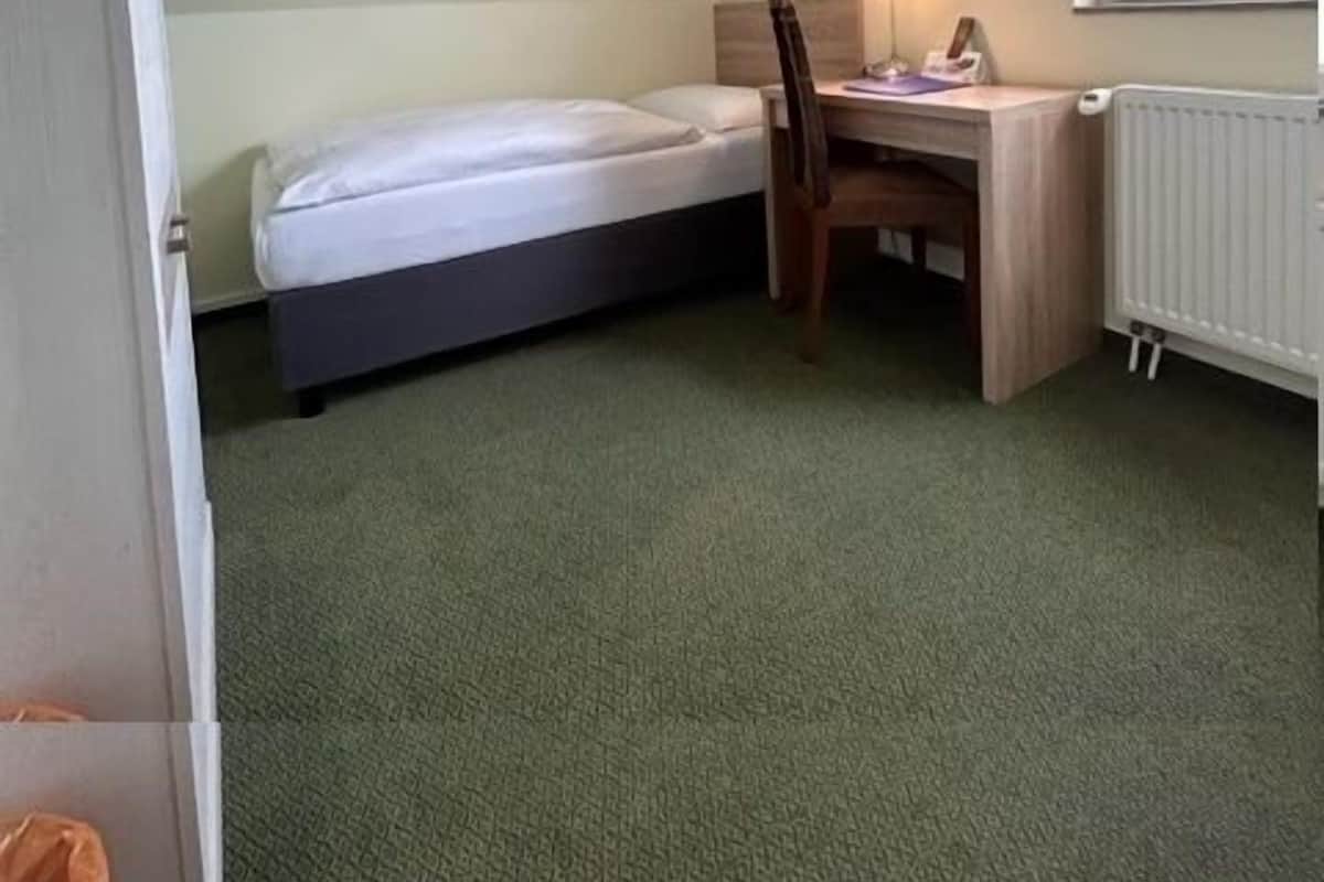 Ein kleines Zimmer mit einem Einzelbett, einem Schreibtisch, einem Stuhl, einer Heizung und einem Fenster mit Blick auf Grünflächen.