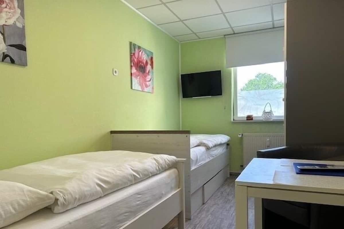 Ein Studentenwohnzimmer mit zwei Einzelbetten, einem Schreibtisch, einem Fernseher und einem Fenster mit Blick auf Grünflächen.