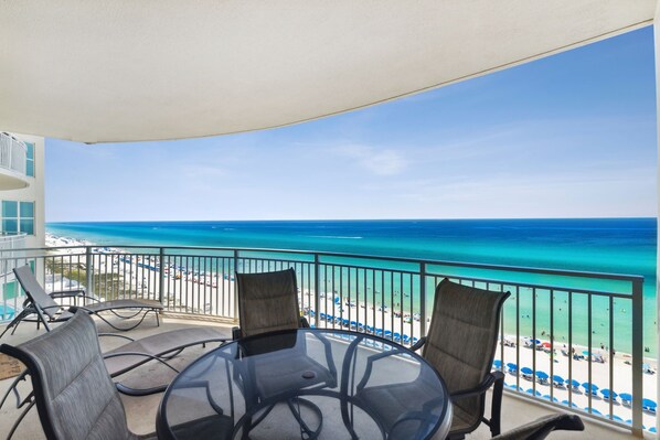 A balcony with a glass table and chairs, overlooking a beach with clear blue water and a sandy shore.